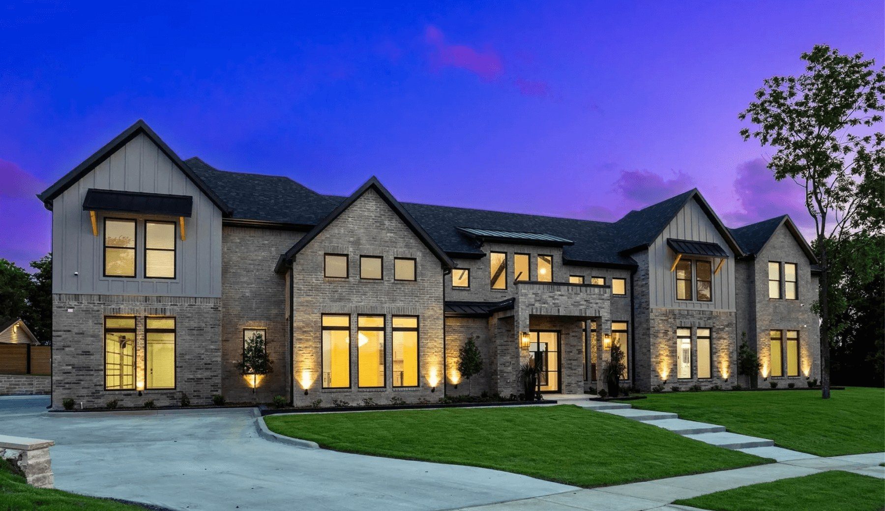 A custom two-story stone and board-and-batten home at dusk, windows lit